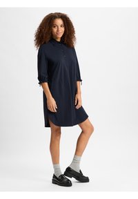 Dunkelblaues Hemdblusenkleid mit hochgekrempelten Ärmeln, seitlichen Schlitzen und einer lockeren Passform. Glatte Textur und knielanges Design. Kombiniert mit schwarzen Schuhen und grauen Socken.