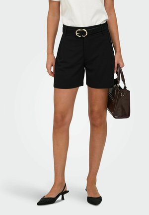 Kvinde iført sorte shorts med bælte, hvid top, sorte kitten heels, der holder en mørkebrun håndtaske mod en ensfarvet baggrund.