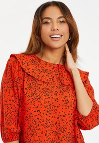 Rode blouse met zwarte luipaardvlekken, voorzien van een gerimpelde kraag, lange pofmouwen en een knoopdetail bij de nek. Soepele textuur.
