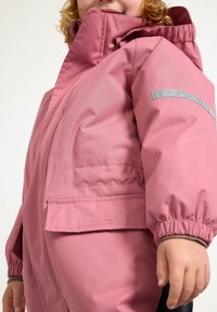 Giacca impermeabile rosa con polsini elasticizzati, cappuccio e dettagli riflettenti sulla manica. Presenta una tasca frontale e una texture liscia.