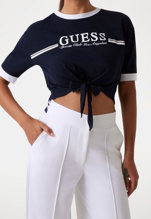 Kvinde iført en marineblå kort cropped t-shirt med knude foran og hvid kant samt hvide højtaljede bukser, hånd i lommen og tatovering på underarmen.