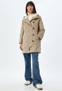 Abrigo impermeable beige con capucha forrada de forro polar, cierres de botón y bolsillos laterales, combinado con vaqueros de mezclilla acampanados y zapatillas blancas.