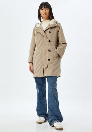 Abrigo impermeable beige con capucha forrada de forro polar, cierres de botón y bolsillos laterales, combinado con vaqueros de mezclilla acampanados y zapatillas blancas.