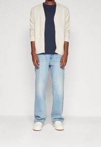 Ljusbeige stickad kofta över en mörk marinblå t-shirt, i kombination med ljusblå jeans och vita sneakers. Koftan har en knappstängning.