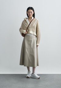 Pull court beige en tricot avec motif texturé, chemise blanche à col, jupe en cuir beige taille haute, et baskets blanches avec accents marron.