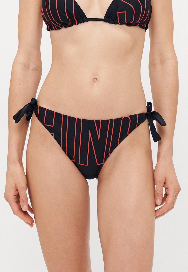 Sieviete valkā melnas bikini apakšdaļas, kurai ir sarkanu kontūru burti un sānu saites, atklājot vidukli, un rokas pret vienkārša fona.