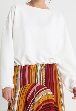 Osoba nosząca biały top z długim rękawem i sznurkiem w talii, połączony z spódnicą z wysokim stanem z pionowymi czerwonymi, pomarańczowymi i białymi paskami.