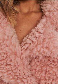 Tejido rosa difuso con una superficie texturizada y esponjosa, que presenta un cuello de chales y un diseño de frente abierto. Textura suave y peluda visible.