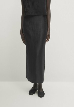 Woman wearing a long, straight black skirt paired with a matching black top and black flat shoes, standing against a light background.