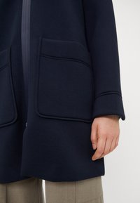 Námořnická modrá vrchní vrstva s hladkým materiálem, dvěma velkými kapsami s kontrastním stehem a minimalistickým designem.
