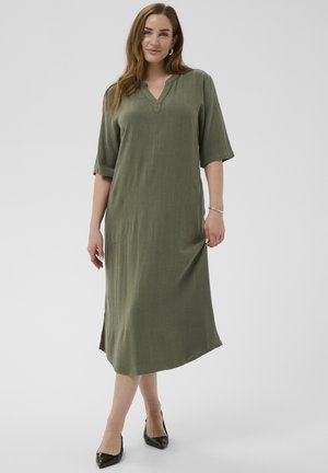 Kobieta ubrana w luźną oliwkowozieloną sukienkę midi z rękawami do łokcia i czarne buty na obcasie z zaostrzonym noskiem, stojąca na jednolitym tle.