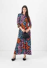 Cvečni dolgi plašč z živahnimi barvami (oranžna, roza, modra) na črnem ozadju, z gumbi spredaj in dolgimi rokavi. Ima majhno črno torbico.