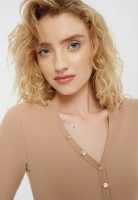 Beige pruhovaný top s výstřihem do V a zlatými knoflíkovými detaily, s přiléhavým střihem a jemnou texturou. Vlasy jsou vlnité a světlé.