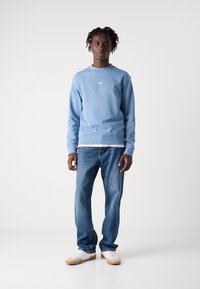 Ανοιχτό μπλε φούτερ με στρογγυλή λαιμόκοψη και λευκό λογότυπο, συνδυασμένο με μπλε τζιν και λευκά αθλητικά παπούτσια με καφέ σόλες.