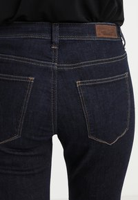 Vaqueros de mezclilla azul oscuro con costuras en contraste y bolsillos traseros, combinados con una blusa negra que se asoma en la cintura.