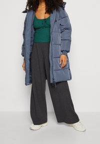 Manteau rembourré bleu foncé avec une coupe mi-longue, associé à un haut côtelé vert et un pantalon gris à jambes larges, avec des baskets blanches.