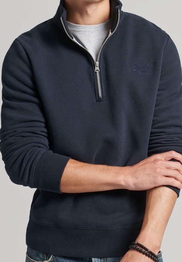 Homme portant un pull bleu marine à fermeture éclair jusqu'à mi-poitrine sur une chemise grise, retroussant une manche, avec des bracelets en perles à un poignet.