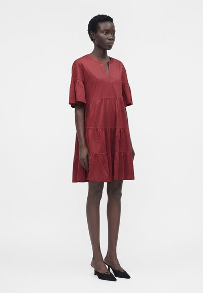 Γυναίκα που στέκεται φορώντας ένα χαλαρό κόκκινο φόρεμα μέχρι το γόνατο με στρωτά βολάν και μαύρα μυτερά τακούνια, σε απλό φωτεινό φόντο.