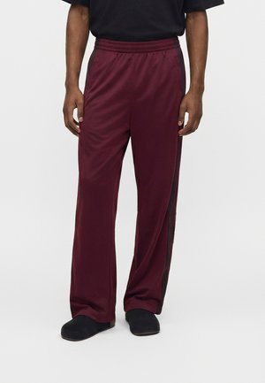Homme portant un pantalon large bordeaux avec des bandes latérales noires et des chaussures noires sans lacets, debout devant un fond uni.