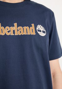 Tumši zils kokvilnas T-krekls ar "Timberland" logo beidzās pelēkā krāsā, virs izliekta līnijas attēlots koks. Īsas piedurknes, apaļa kakla izgriezums.