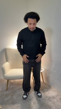 Bărbat cu păr creț, purtând un hanorac negru și blugi închiși la culoare, stând lângă un fotoliu alb și o masă rotundă mică, într-o cameră luminată delicat.