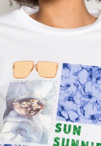 Camiseta blanca con un diseño gráfico que presenta unas gafas de sol naranjas, un vaso floral, patrones azules y la frase "SUN SUNSHINE" en cristales de roca verdes.