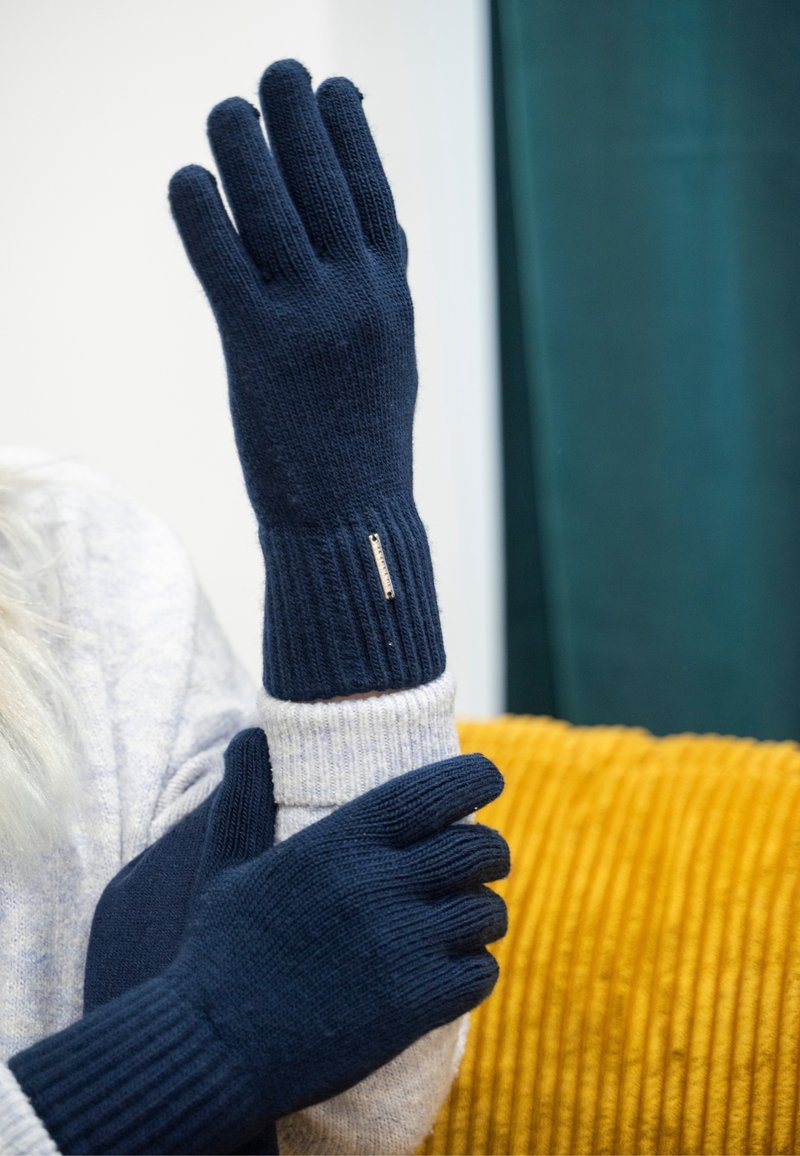 Marineblaue Strickhandschuhe mit gerippten Bündchen und Fingeröffnungen. Sie verfügen über eine erhabene Textur und ein kleines metallisches Akzent nahe dem Handgelenk.