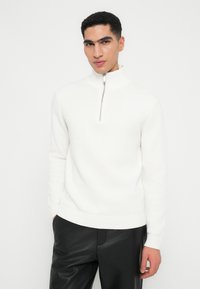 Junger Mann in einem weißen Strickpullover mit einem halben Reißverschluss und schwarzen Lederhosen, der mit einer Hand in seiner Tasche steht.