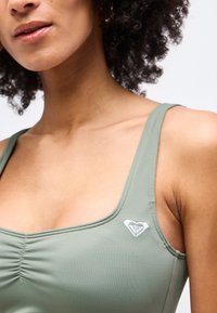 Γυναίκα με σγουρά μαλλιά που φοράει ανοιχτό πράσινο τοπ με ένα μικρό λευκό λογότυπο σε σχήμα καρδιάς στο στήθος.