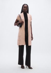 Μακριά, ανοιχτού μπροστινού μέρους ζακέτα σε ανοιχτό ροζ πλεκτό με υφή μοτίβων. Φορεμένη πάνω από μαύρο εφαρμοστό τοπ και συνδυασμένη με μαύρο παντελόνι φαρδιάς γραμμής.