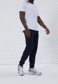Witte katoenen T-shirt met korte mouwen, gecombineerd met marineblauwe joggingbroek. Zilveren en gouden hardloopschoenen van mesh en rubber.