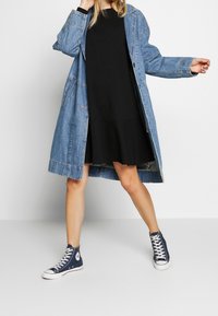 Veste longue en denim, bleu clair, avec boutons sur le devant et poches latérales, portée sur une robe noire. Des baskets montantes bleu marine complètent la tenue.