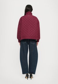Maroon quilted jacket with high collar, ribbed cuffs, and a boxy fit; paired with wide-leg blue jeans and black heeled shoes.