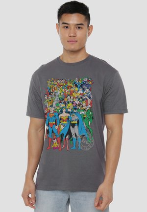 Young man wearing a gray t-shirt with a colorful print of various classic DC Comics superheroes standing together.