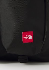 Zbliżenie na czarny plecak z czerwoną metką z logo "THE NORTH FACE". Tkanina ma teksturowany wzór.