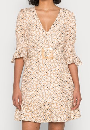 Mujer que lleva un vestido beige con estampado floral, escote en V, mangas abullonadas, bajo con volantes y un cinturón a juego con una hebilla cuadrada.