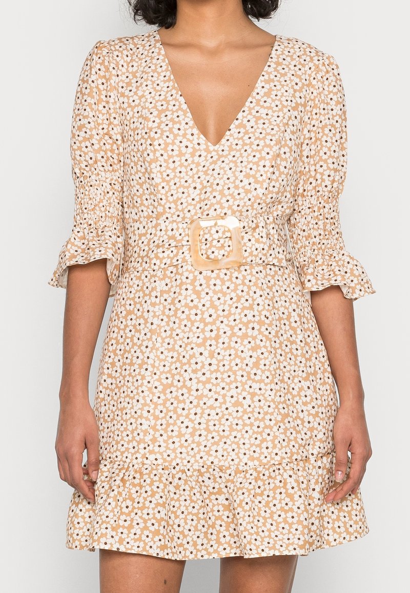 Femme portant une robe beige à fleurs avec un col en V, des manches bouffantes, un ourlet à volants et une ceinture assortie avec une boucle carrée.