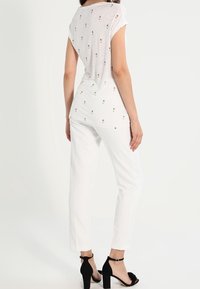 Top bianco a maniche corte con una vestibilità ampia, decorato con piccoli motivi floreali. Abbinato a pantaloni bianchi a vita alta e sandali neri con tacco.