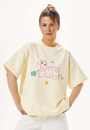 Kobieta z włosami związanymi w kok, ubrana w jasnopastelow żółtą luźną koszulkę z kolorowym napisem "Moms Club" oraz małymi ilustracjami samochodu, kwiatu, gwiazd i ptaka.