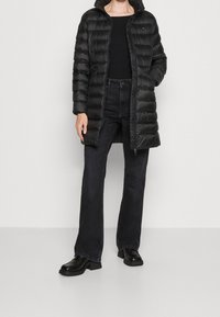 Abrigo acolchado negro con patrón de acolchado horizontal, cierre con cremallera y capucha. Llevado sobre una camiseta negra y jeans oscuros, combinado con botas negras gruesas.