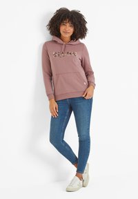 Dusty rose hoodie with "Bloom" text in floral pattern, front pouch pocket, ribbed cuffs, paired with blue skinny jeans and white sneakers.