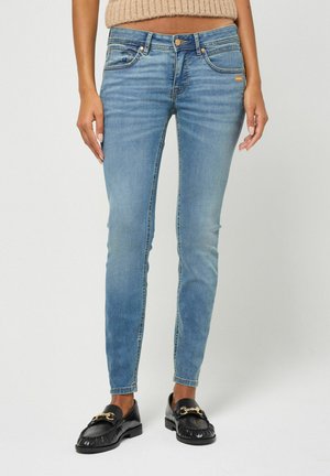 Frau trägt hellblaue Skinny Jeans und schwarze Loafers mit goldener Kettendetail, steht vor einem schlichten Hintergrund.