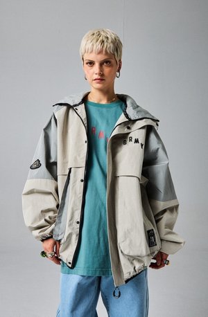Chaqueta ligera en bloques de color beige y gris, con capucha, grandes bolsillos y un parche del logo en la manga; llevada sobre una camisa color teal.