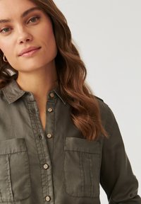 Camisa abotonada de color verde oliva, confeccionada en una tela suave, con cuello, dos bolsillos en el pecho y botones marrones.