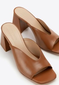 Mules de tacón de cuero marrón con una abertura amplia y punta cuadrada, con un tacón bloque y un acabado suave. Hechos a mano en Italia.
