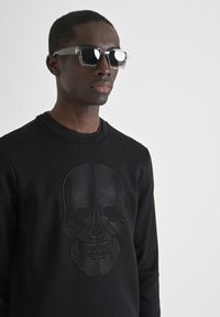 Sudadera negra con un diseño de calavera en relieve en un patrón de líneas onduladas. Usuario usando gafas de sol con montura transparente y lentes oscuros.