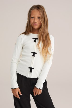 Haut blanc à manches longues avec une texture côtelée, doté de quatre accents de nœud noir et d'un ourlet en forme de vagues. Associé à un pantalon noir, mettant en avant le contraste.