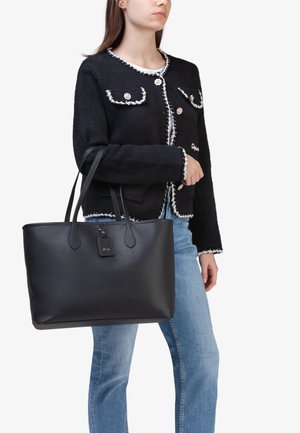 Borsa a mano in pelle nera con finitura texturizzata, due manici superiori e un'etichetta removibile. Abbinata a una giacca nera e jeans blu.