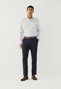 Bărbat îmbrăcat cu o cămașă albă cu flori, mânecă lungă, pantaloni bleumarin, curea maro și pantofi eleganți maro, stând cu o mână în buzunar pe un fundal alb.