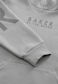 Gaišs pelēks kapucis ar ribotām aprocēm, priekšējo kabatu un reljefa logo tekstu "BAKER by TED BAKER" uz krūtīm un piedurknes.